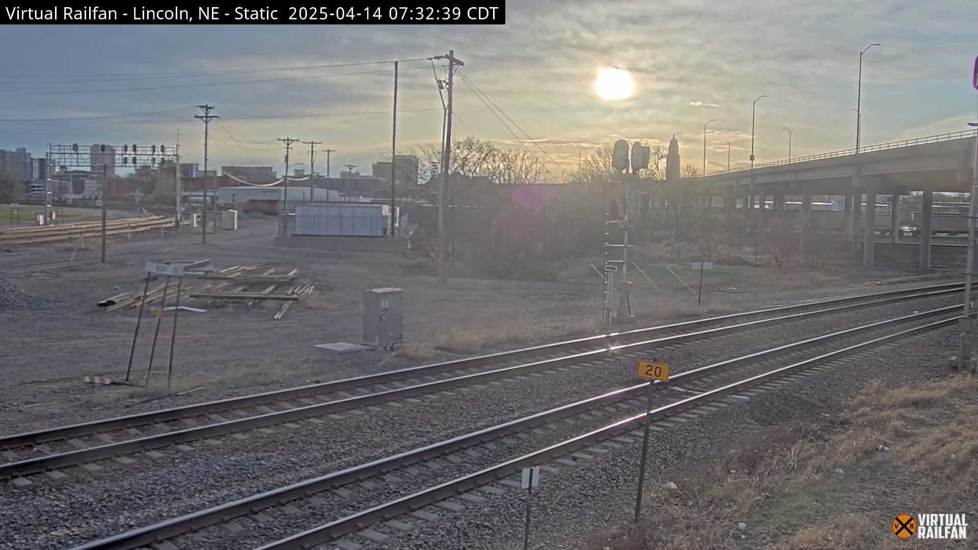 A sunny, partly cloudy day reveals a view of railroad tracks, a highway overpass, and a mostly barren landscape with some sparse vegetation and construction materials.