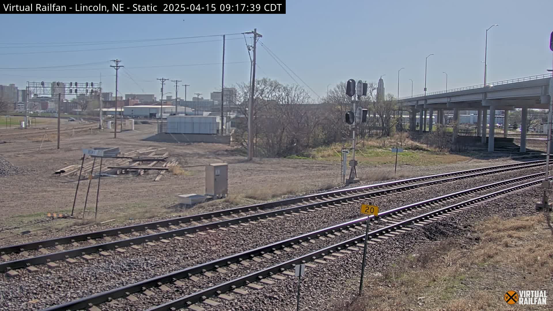 Under a clear sky, railroad tracks extend into the distance, passing under a large overpass and towards a city skyline.
