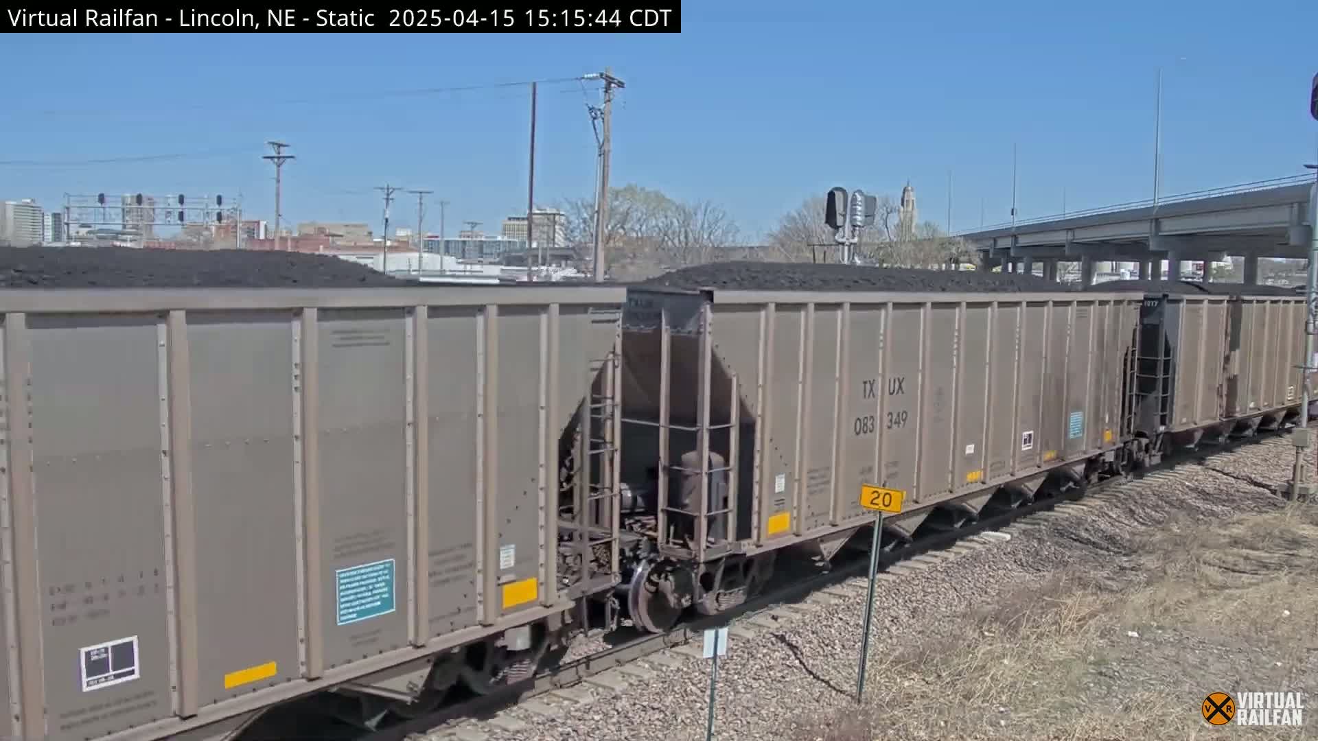 A long freight train carrying coal is visible on a sunny day, passing under an overpass.