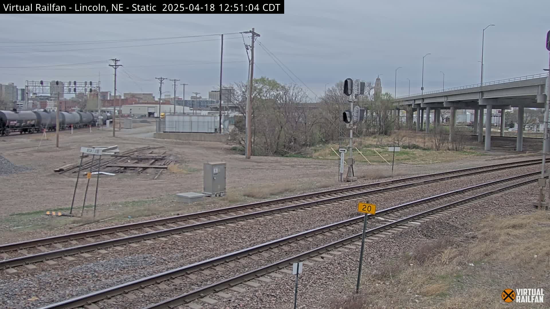 A view of train tracks under an overpass on an overcast day, with a city skyline in the background and a few dormant trees in the foreground.