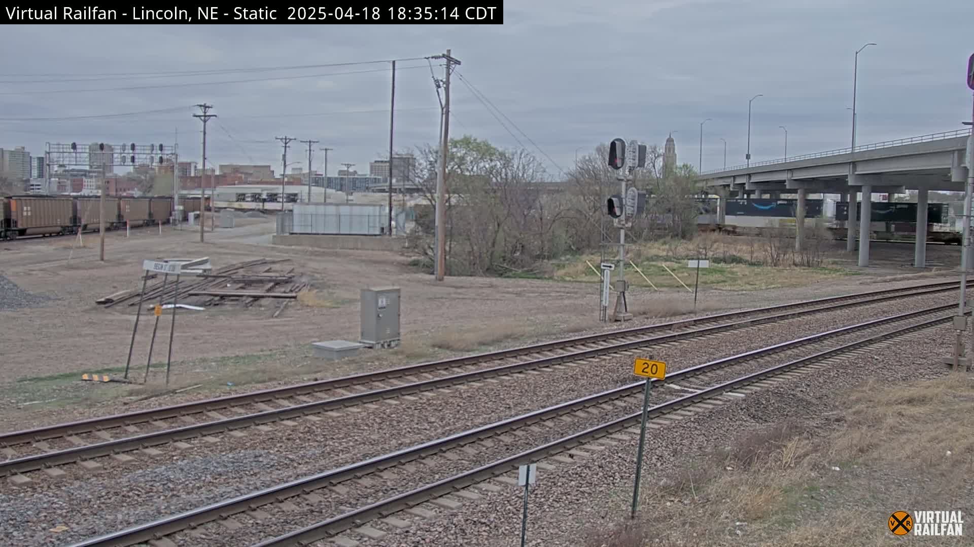 A view of train tracks under an overpass on an overcast day, with a city skyline in the background and several freight cars visible.