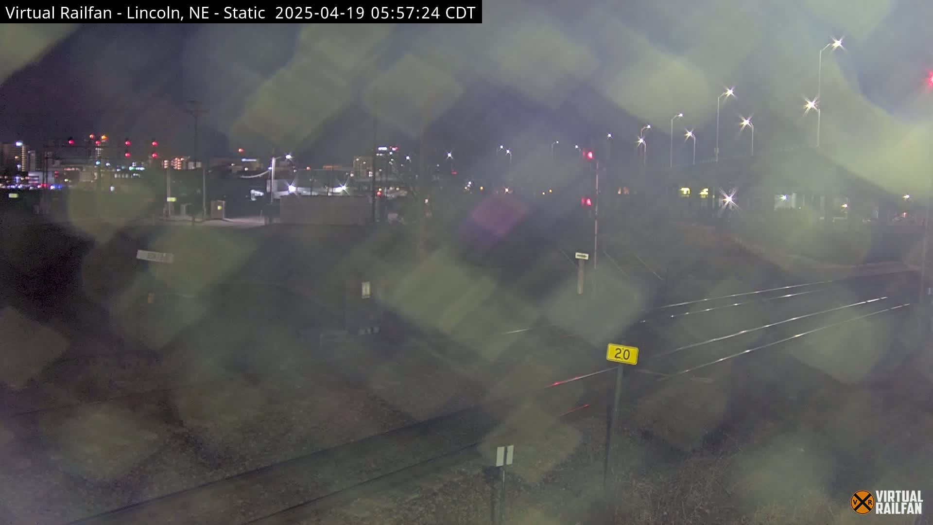 A nighttime view of railroad tracks and a city skyline in the distance, with light rain or mist obscuring the view.