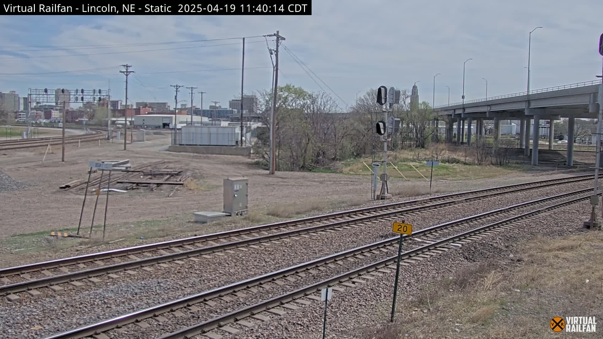 A view of railroad tracks under a partly cloudy sky, with a city skyline in the background and a highway overpass in the mid-ground.