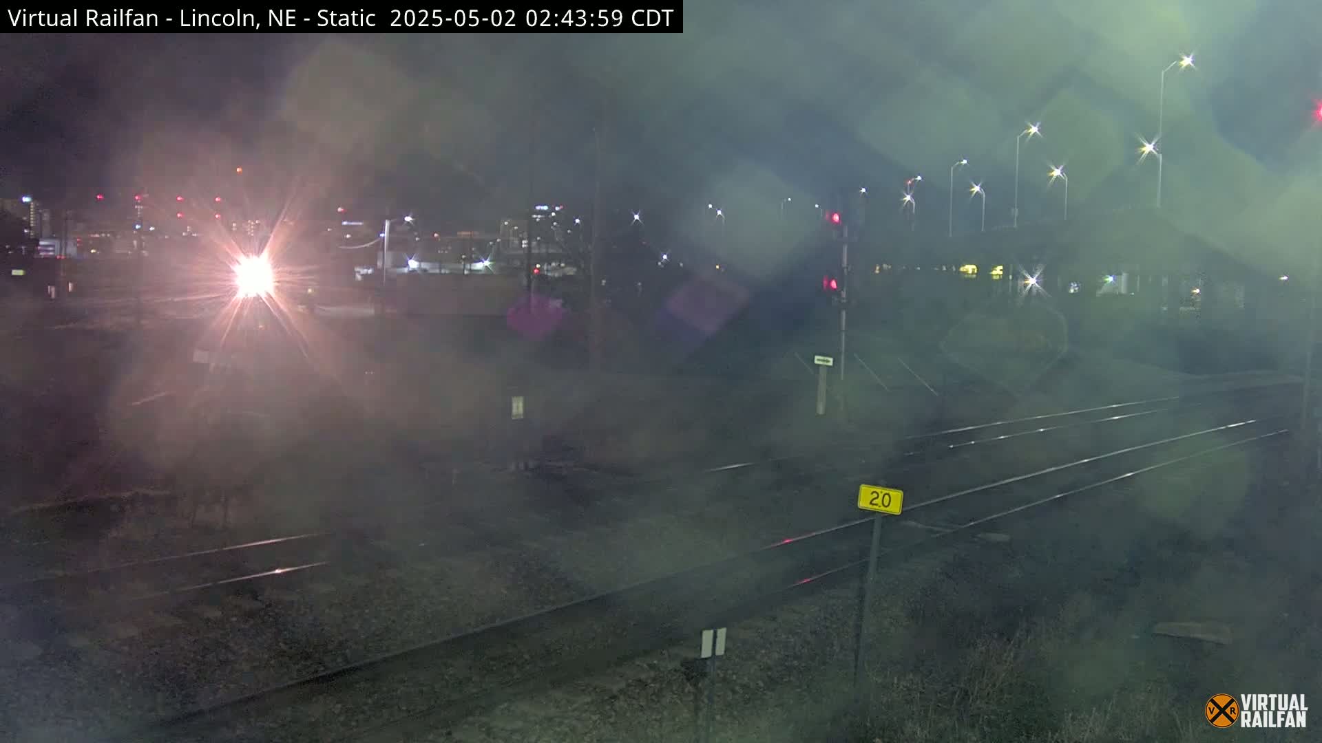 A nighttime view of train tracks with a bright headlight in the distance and hazy weather conditions.