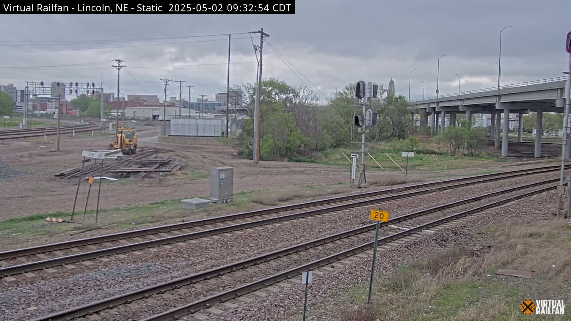 Under an overcast sky, railroad tracks run through a mostly barren landscape with some vegetation, a small yellow piece of construction equipment, and a distant city skyline visible.