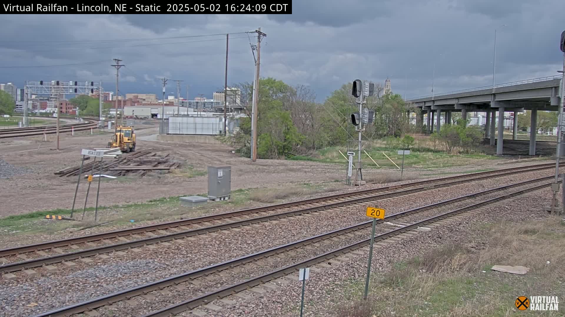 A view of railroad tracks under an overcast sky, with a yellow piece of construction equipment near a pile of railway ties and a highway overpass in the background.