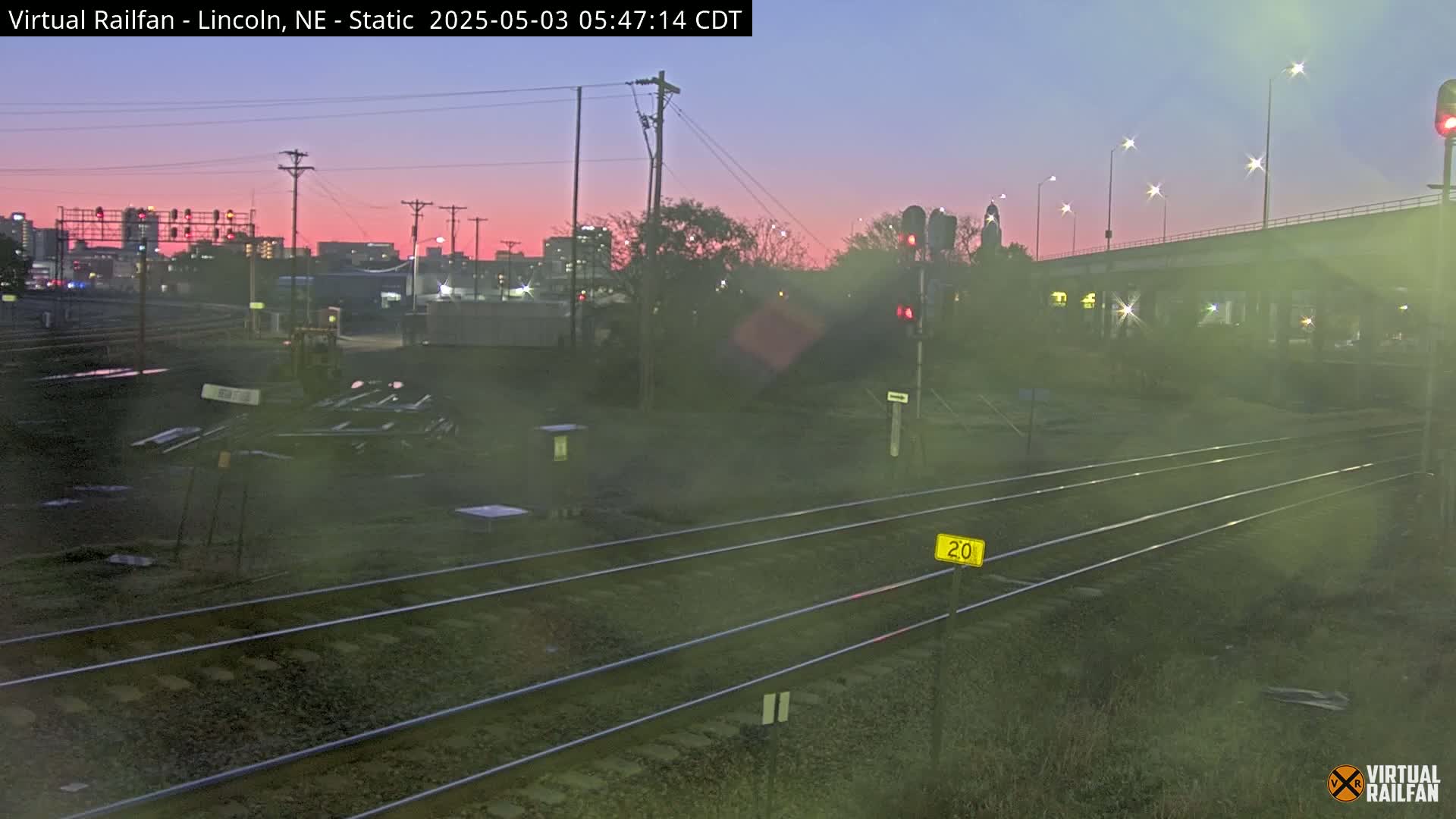 A twilight view of railroad tracks, with a city skyline visible in the distance under a pinkish-purple sky.