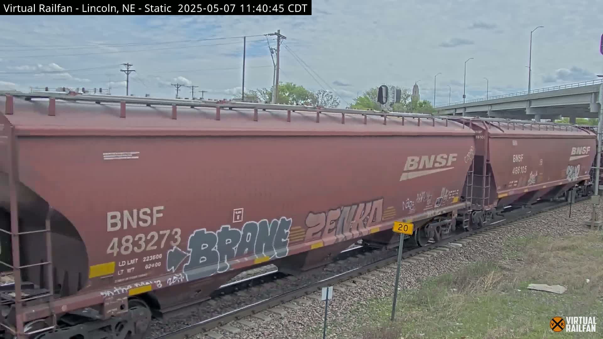 A long reddish-brown freight train with graffiti on its cars moves along railroad tracks under a partly cloudy sky.