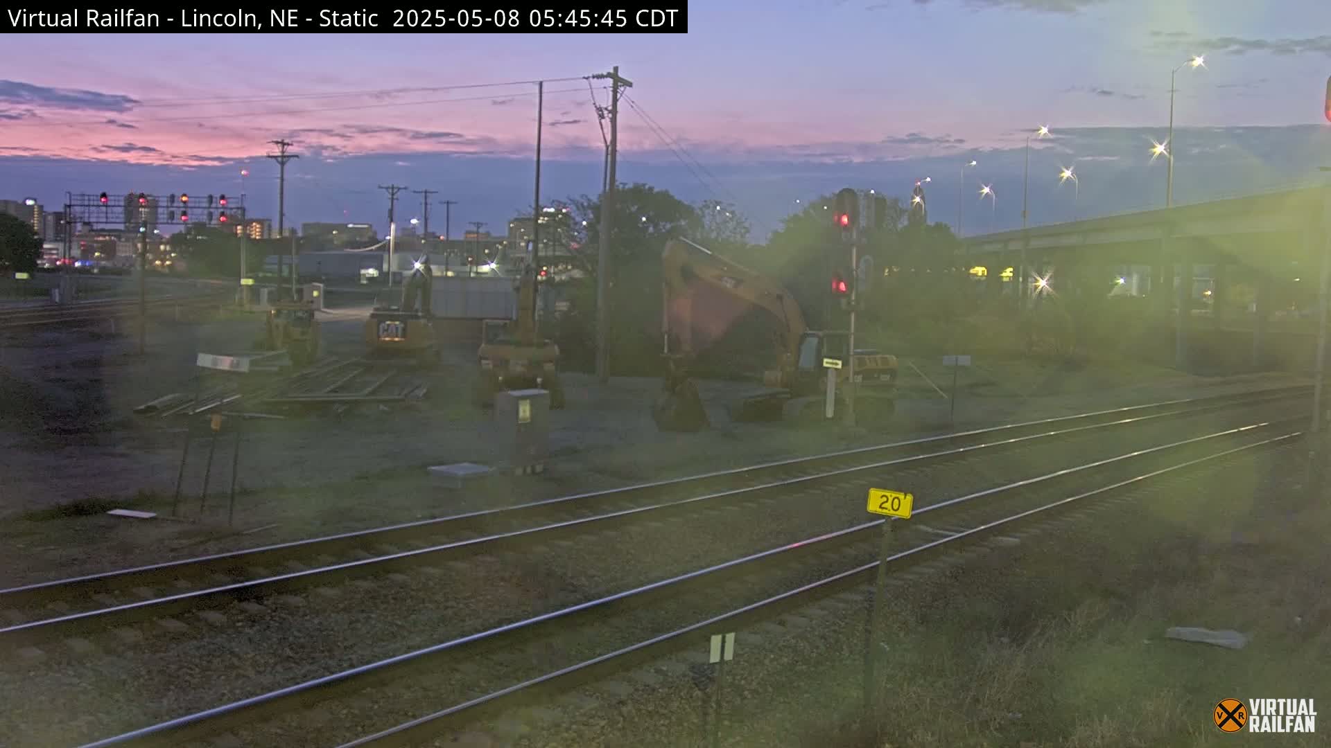 Under a twilight sky, construction equipment sits near railroad tracks in a city setting.