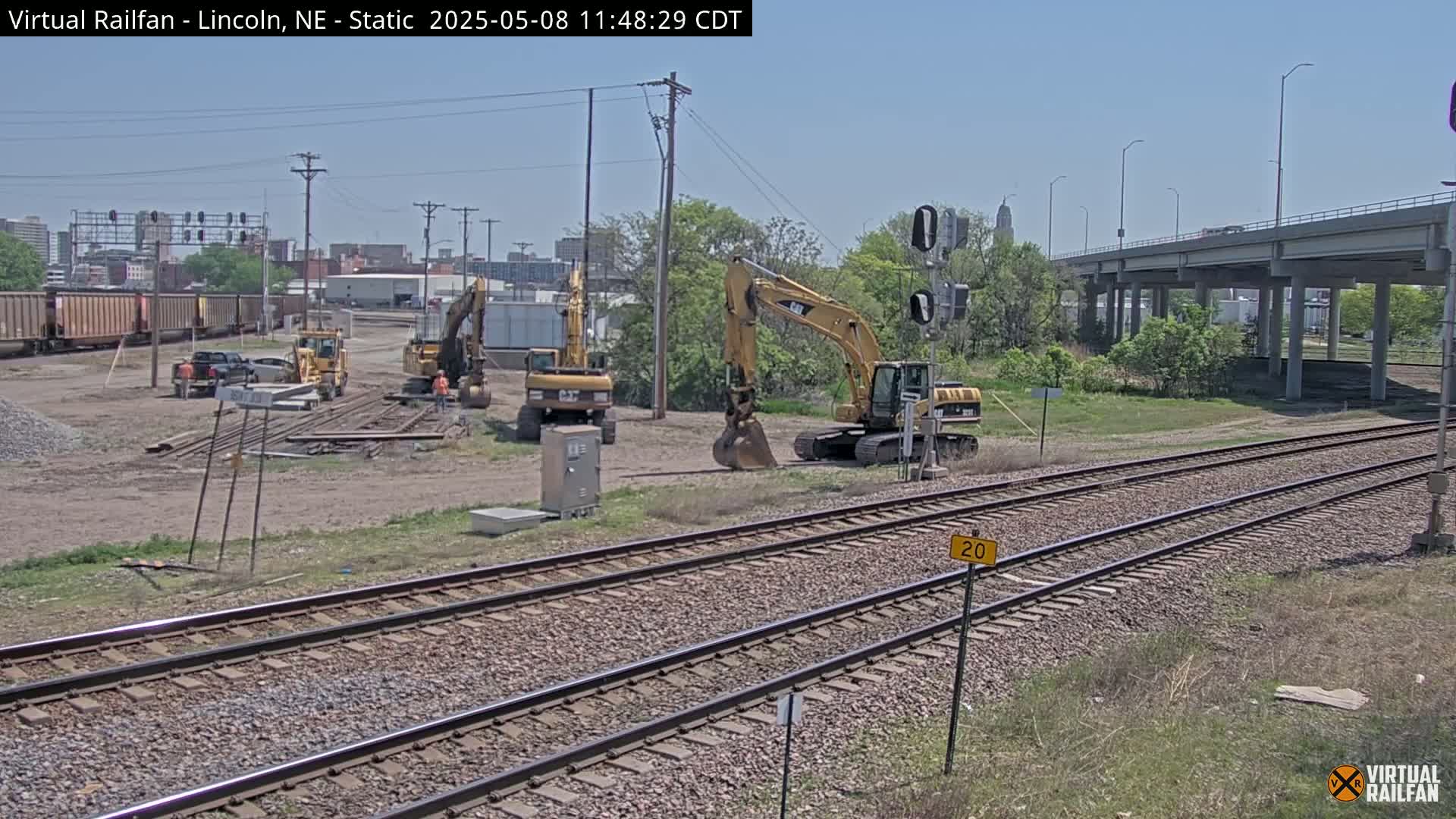 Under a clear sky, several yellow excavators are parked near railroad tracks undergoing construction, with a train car visible in the background.