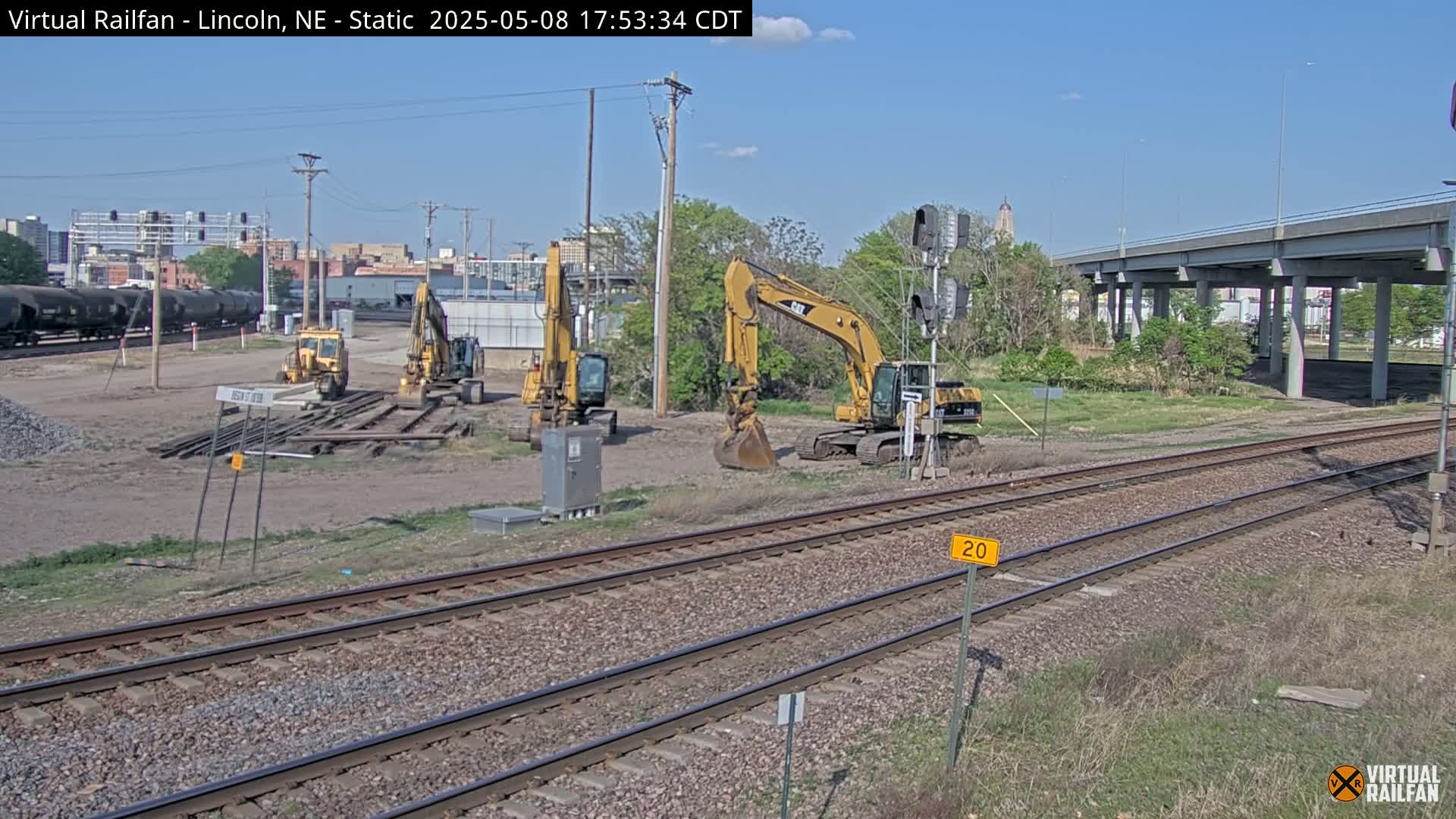 Under a clear blue sky, several yellow excavators sit on a construction site near railroad tracks and a highway overpass.