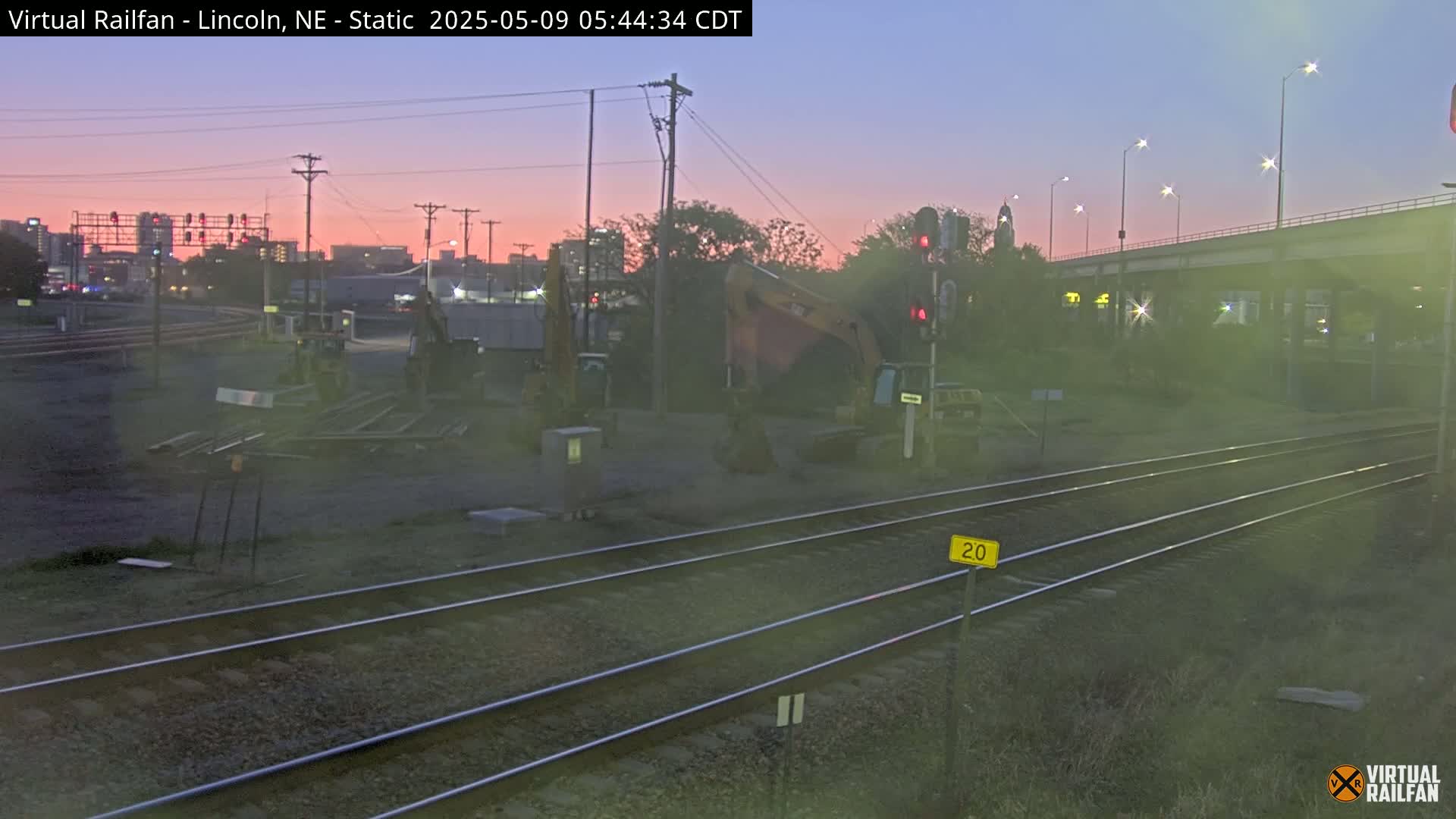 Under a twilight sky, construction equipment sits near railroad tracks in a dimly lit urban area.