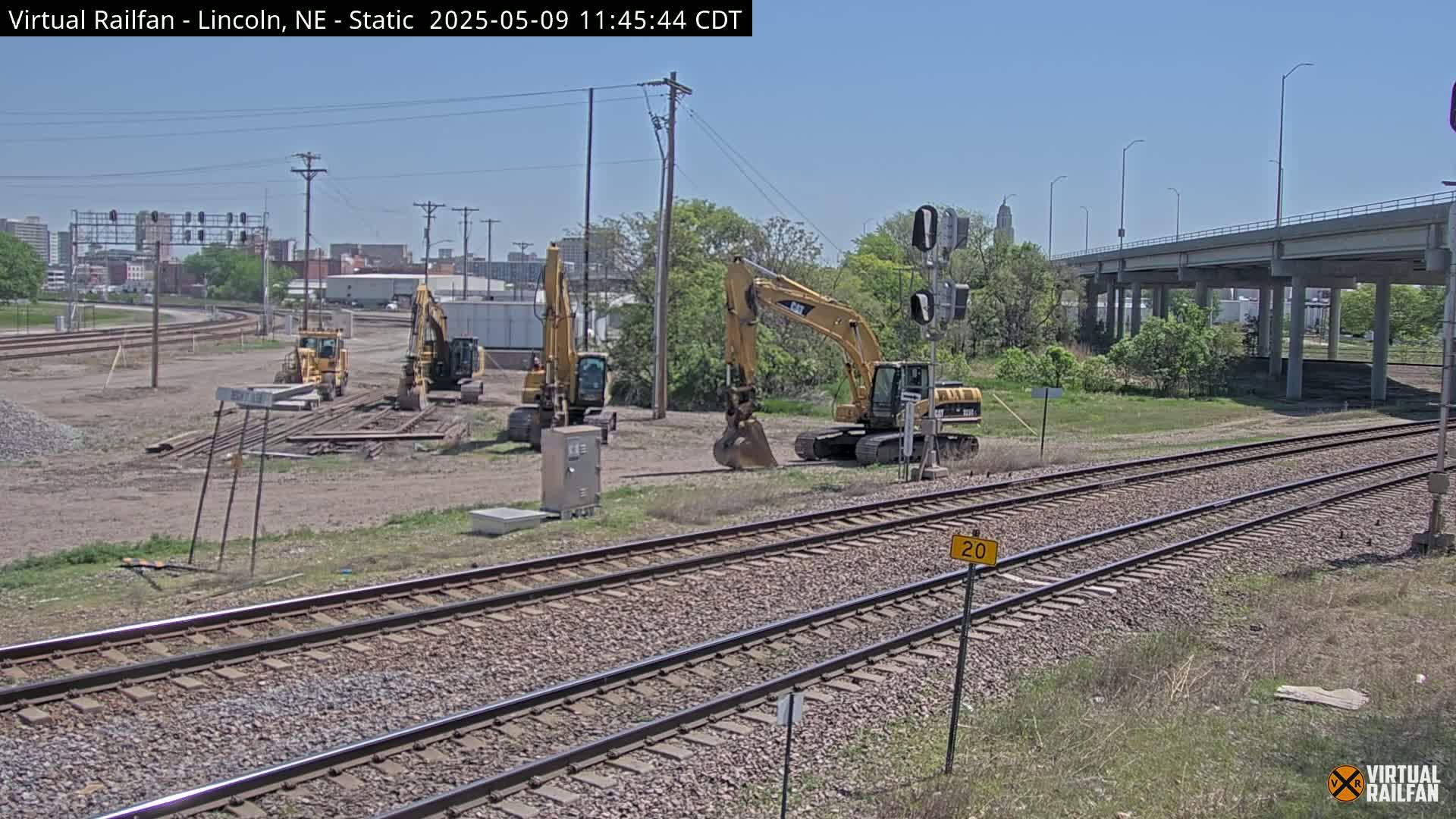 Under a clear blue sky, several yellow excavators sit on a dirt construction site next to railroad tracks that run beneath a highway overpass.