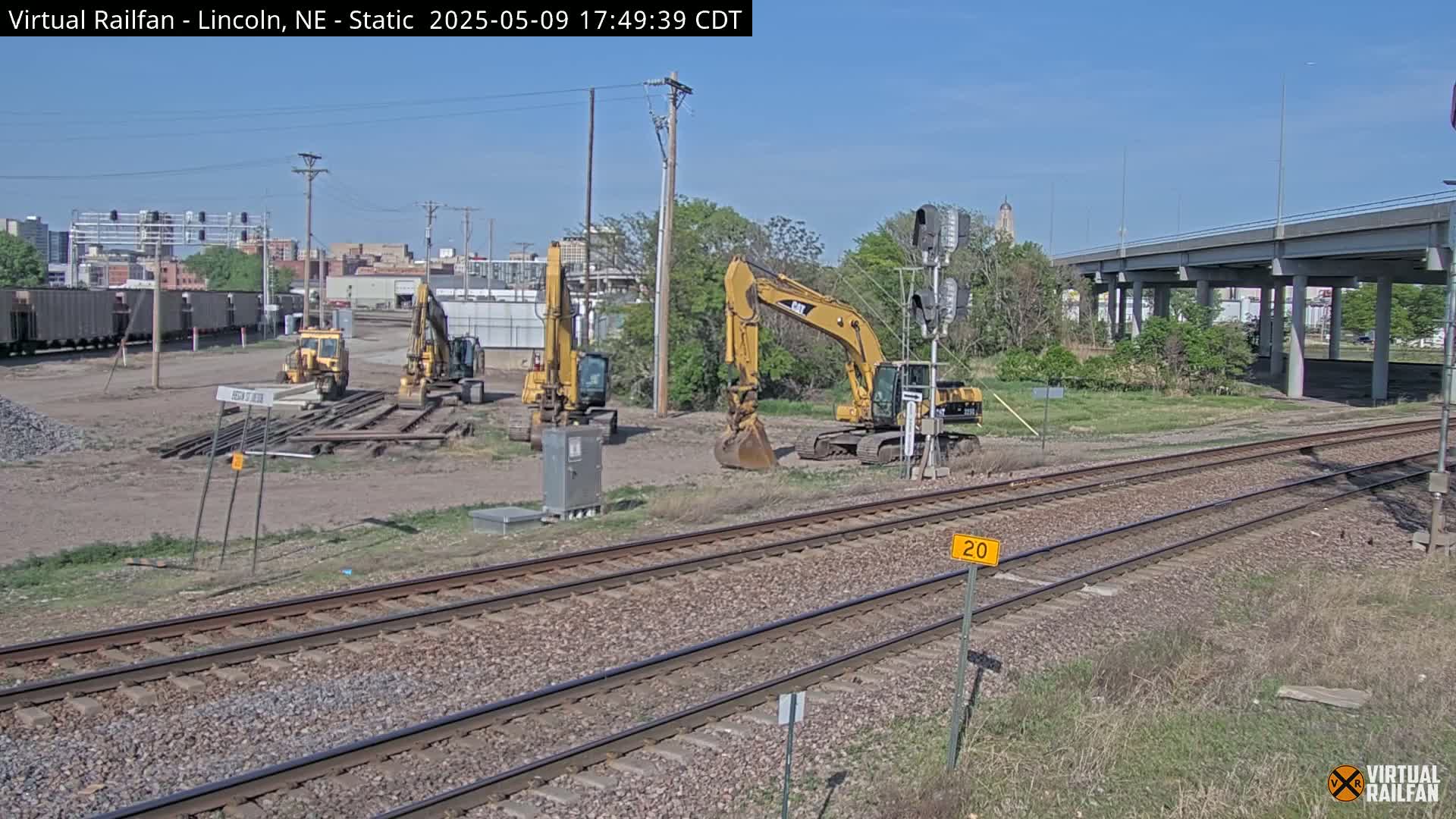 Several yellow excavators are parked near railroad tracks on a sunny day, with a highway overpass visible in the background.