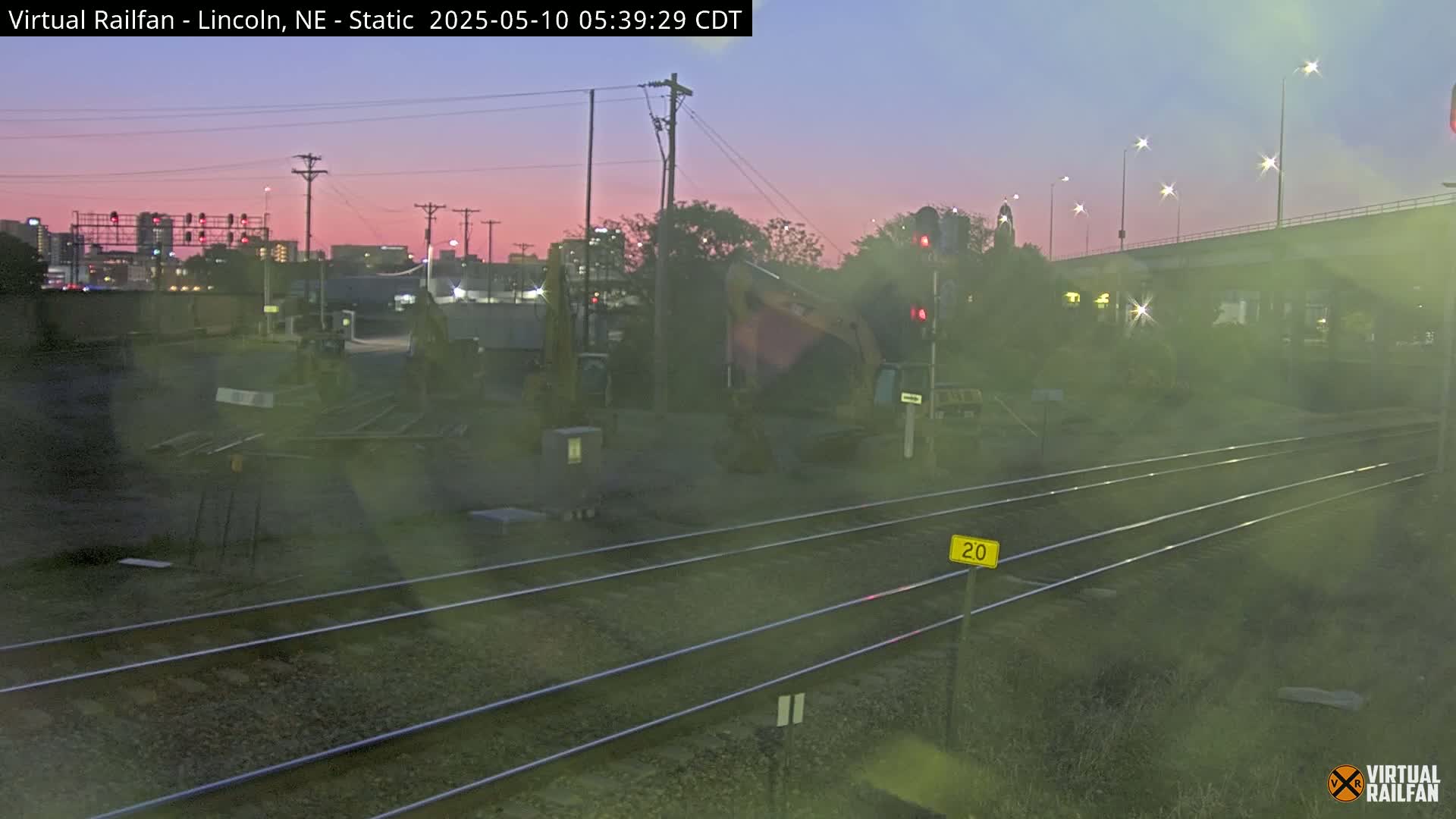 A twilight view of railroad tracks near a city skyline, with a piece of heavy machinery visible, under a dusky pink sky.