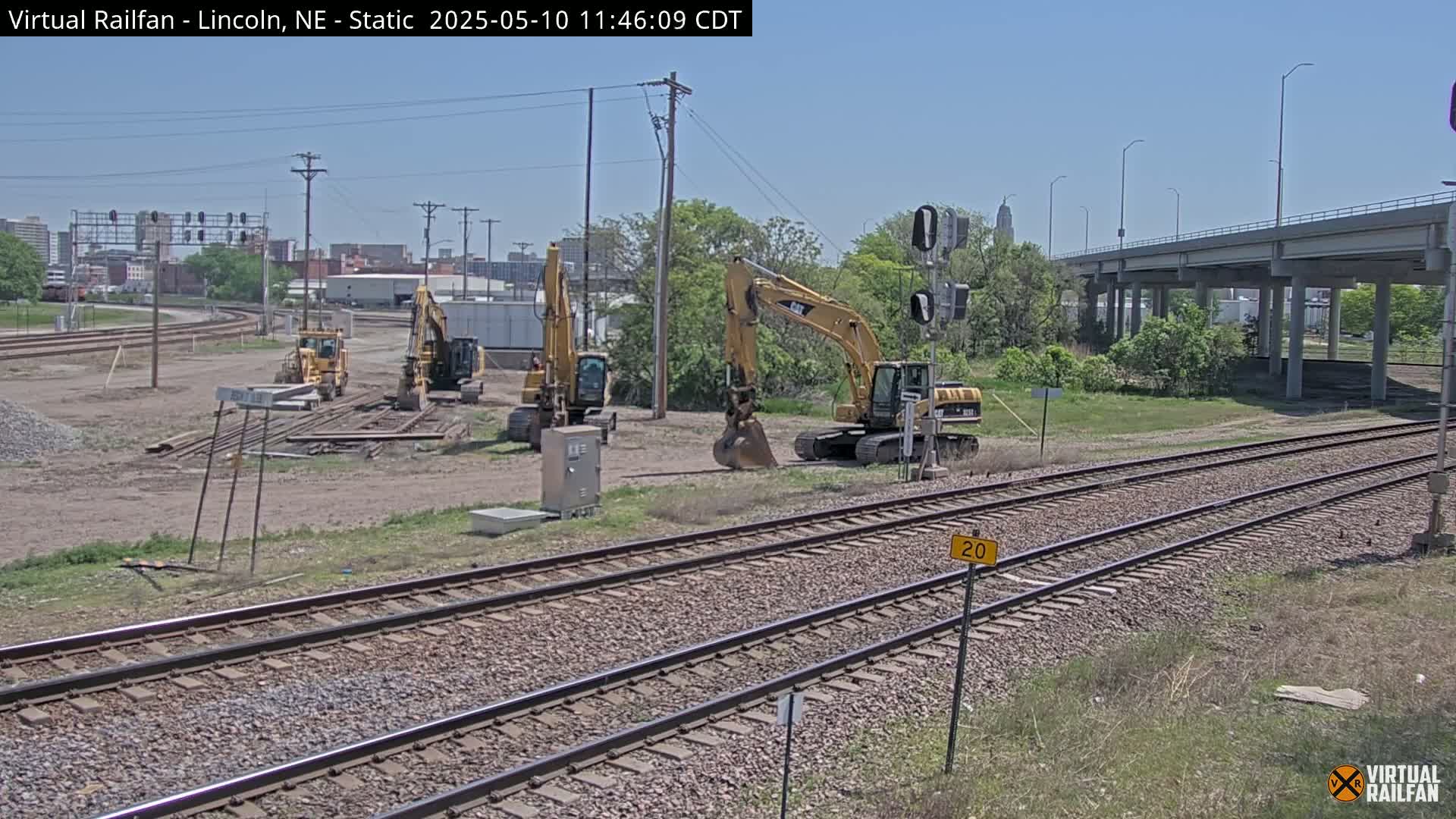 Several yellow excavators are parked near a railway track under a sunny sky.