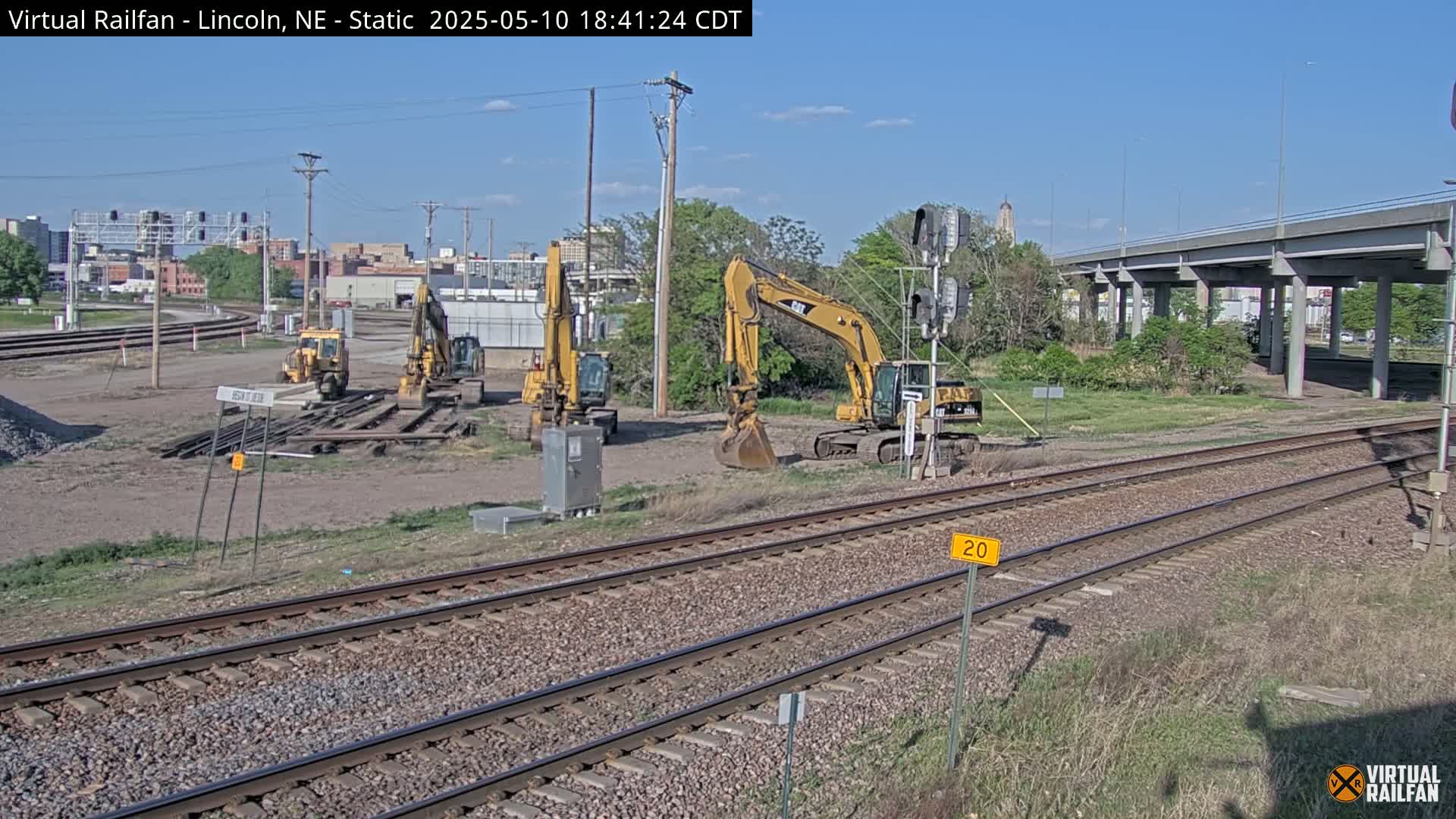 Several yellow excavators are parked near a railway under a partly sunny sky.