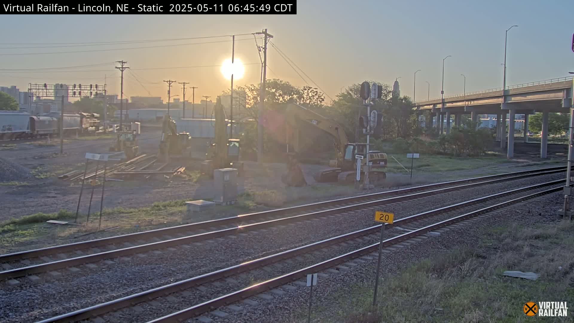 Construction equipment sits near railroad tracks at sunrise under a clear sky.