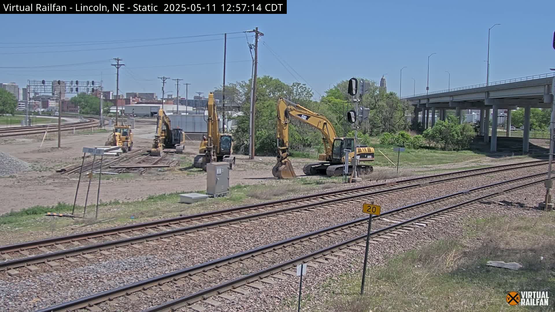 Several yellow construction excavators are parked near railroad tracks on a sunny day.