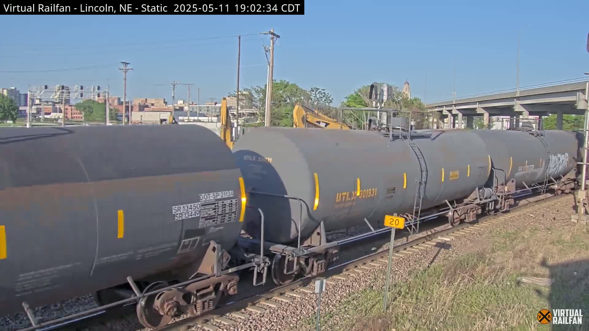 A long freight train of gray tanker cars sits on railroad tracks under a clear, sunny sky.