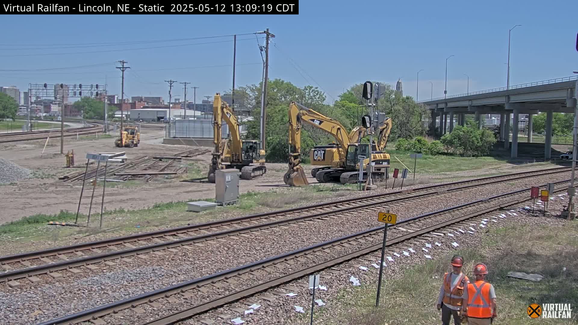 Under a clear sky, two large yellow excavators are parked near railroad tracks next to a construction site, where two workers in bright safety vests stand observing the area.