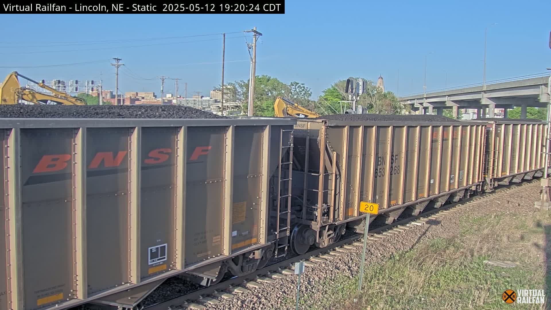 Under a clear blue sky, a long freight train carrying coal is being loaded by two excavators.