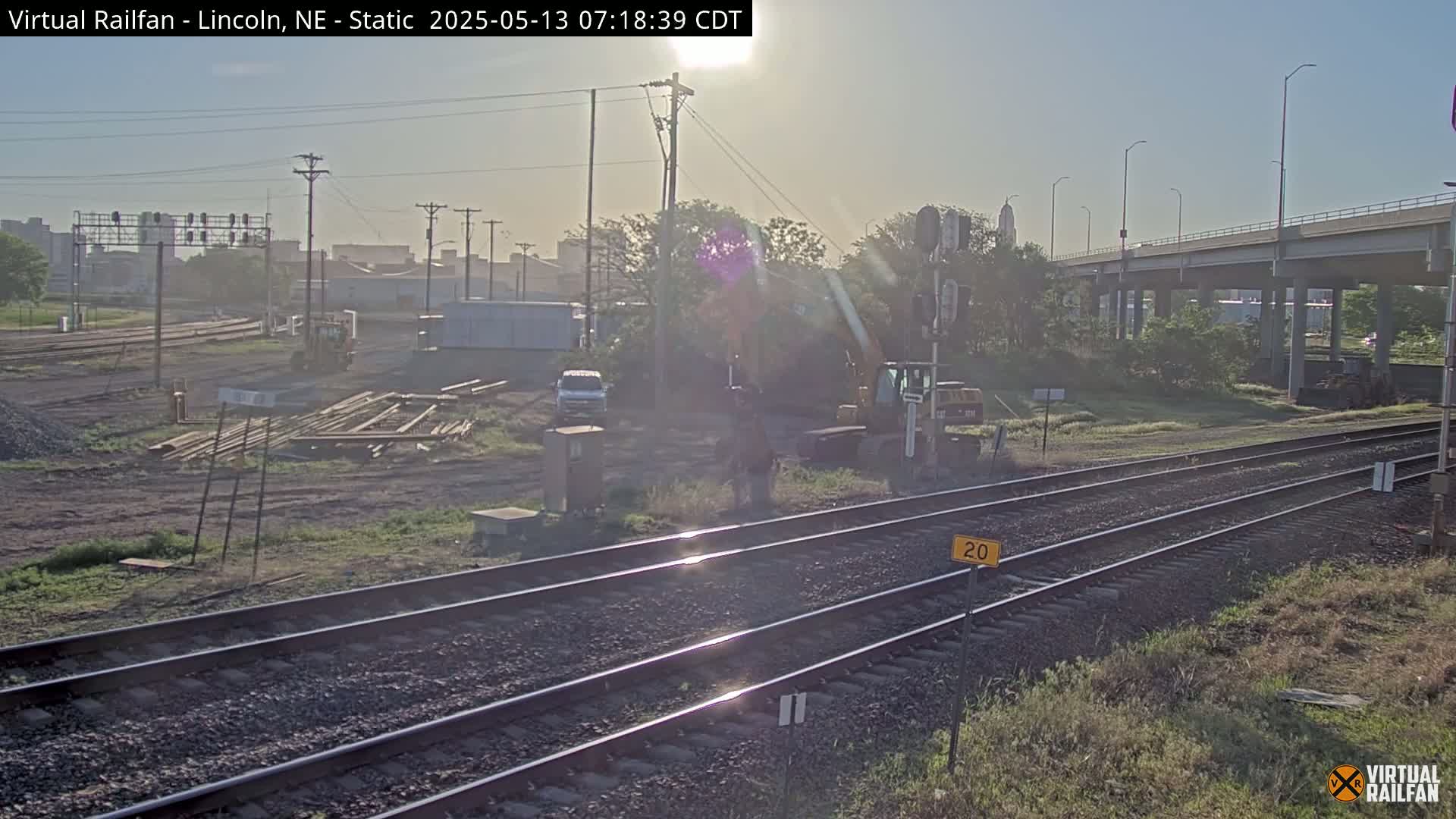 A sunny morning view of railroad tracks, construction materials, and heavy machinery near a highway overpass.