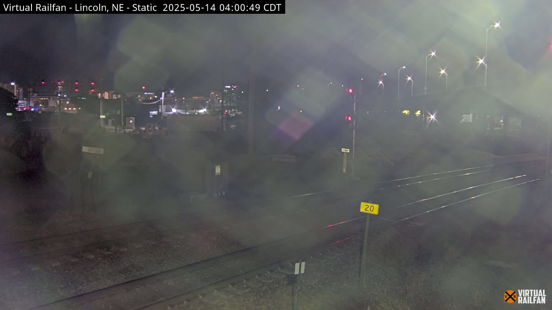 A nighttime, hazy view of railroad tracks with a blurry city background and streetlights.