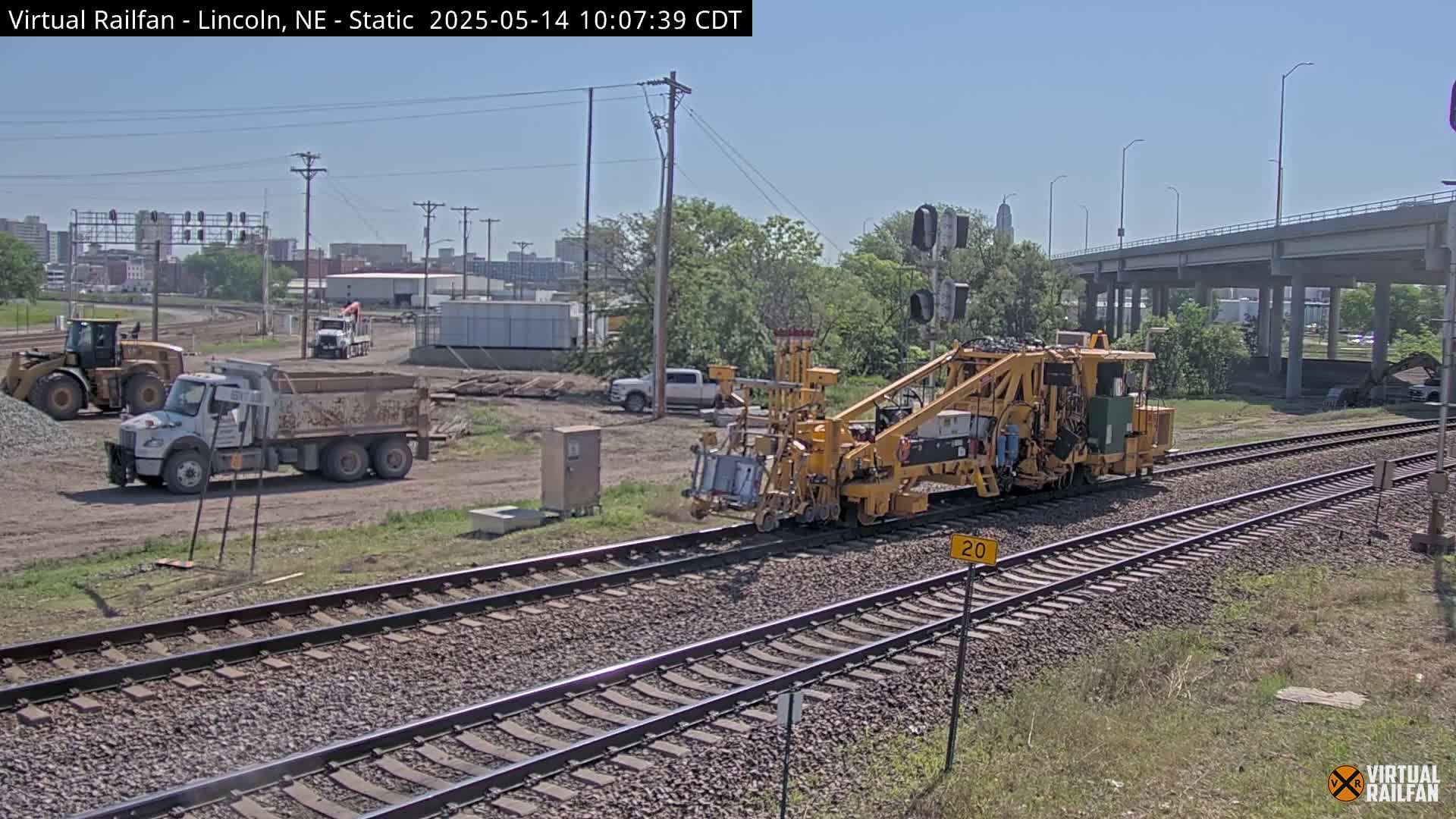 A yellow railway maintenance vehicle travels along train tracks next to a construction site under a sunny sky, with dump trucks and other heavy machinery visible.