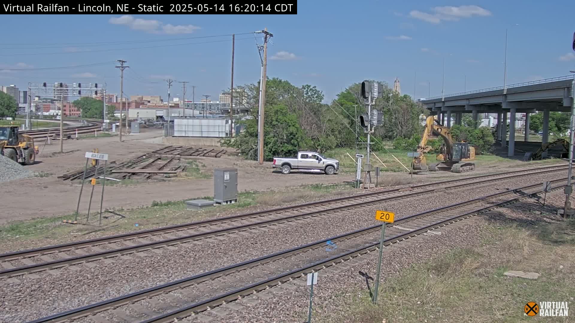 A sunny day reveals a construction site near railroad tracks, featuring several pieces of heavy machinery, a pickup truck, and a city skyline in the background.