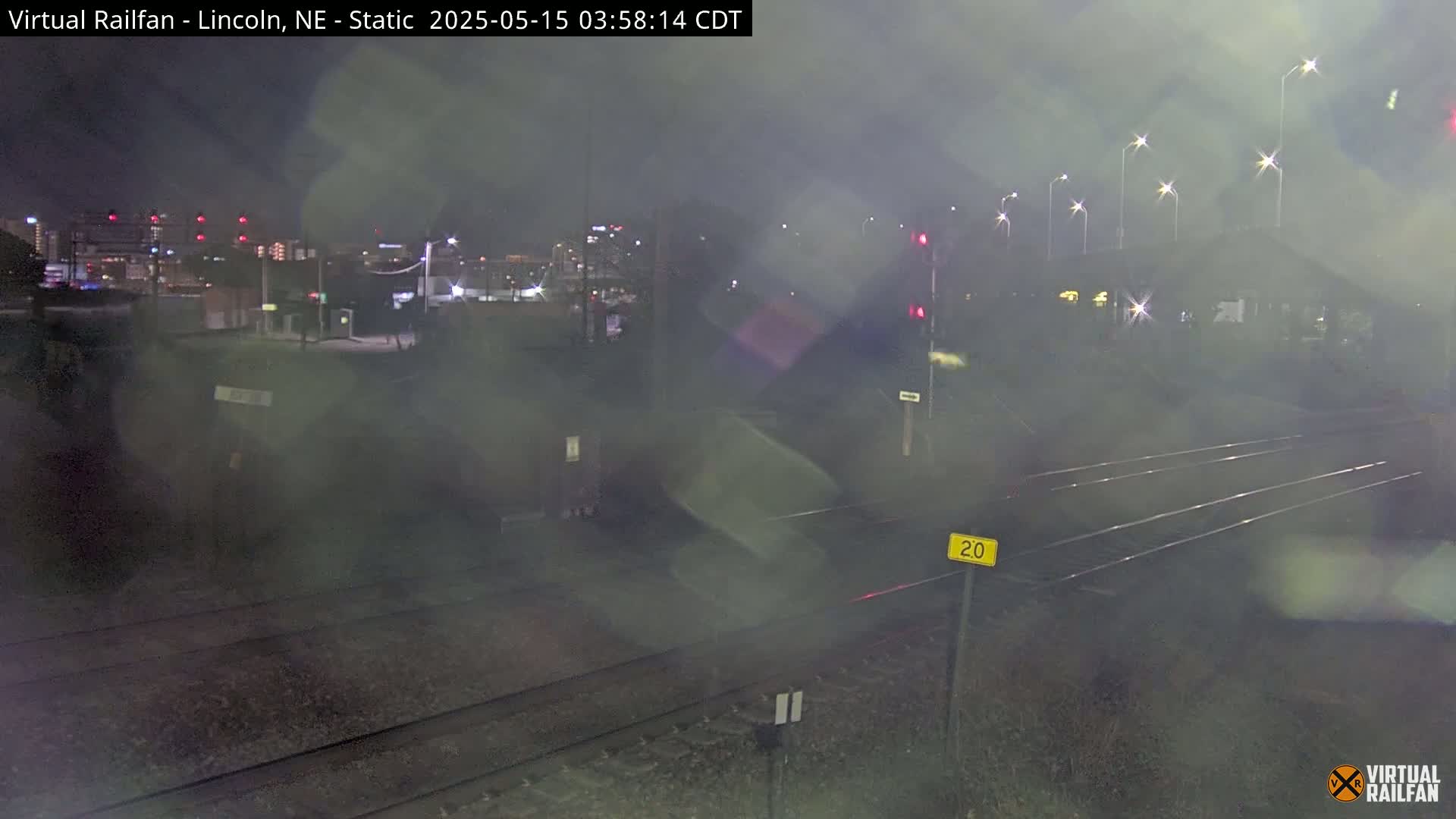 A nighttime view of a railroad crossing with a hazy or foggy atmosphere, showing tracks and distant city lights.