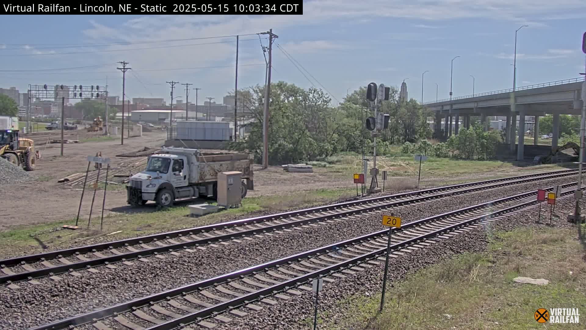 A partly cloudy day shows a construction site near railroad tracks, with a dump truck parked nearby, and a highway overpass in the background.