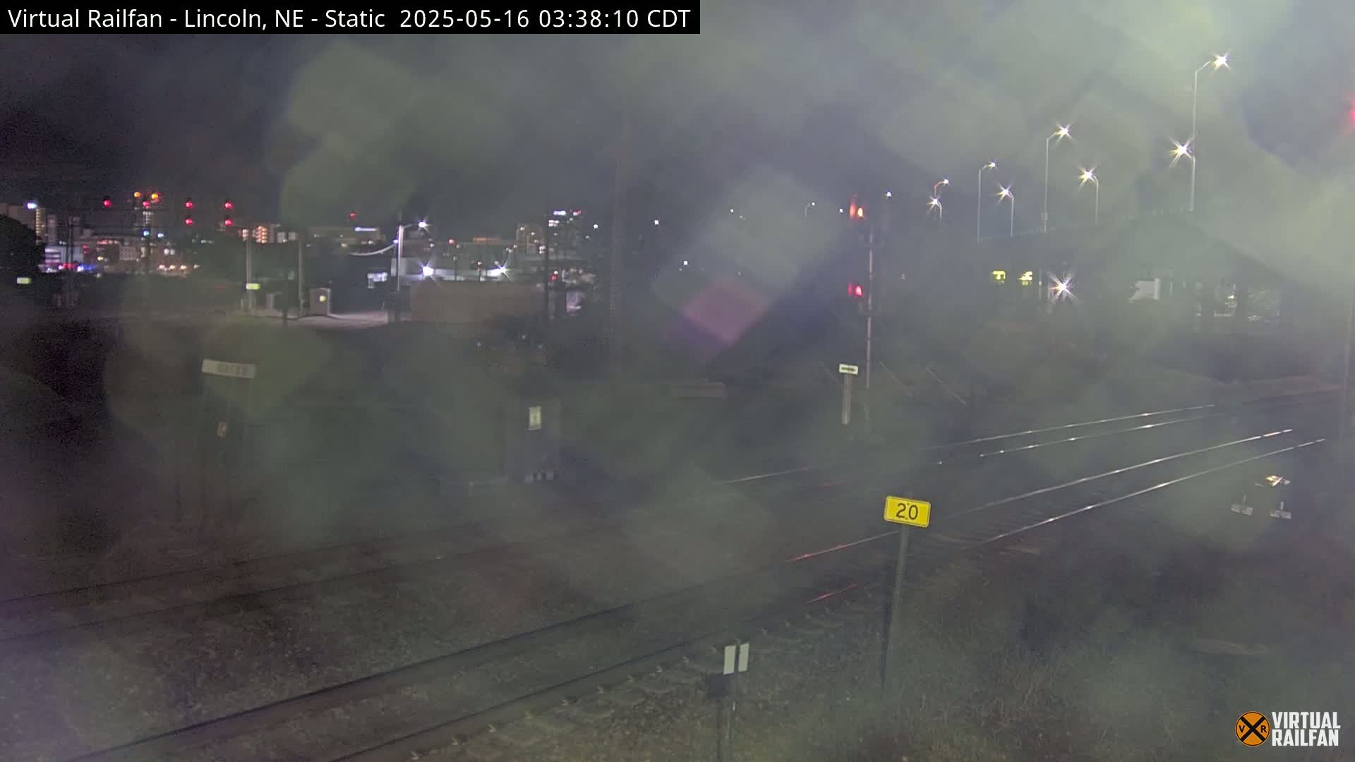 A nighttime view of railroad tracks in foggy conditions, with a distant city skyline illuminated by lights.