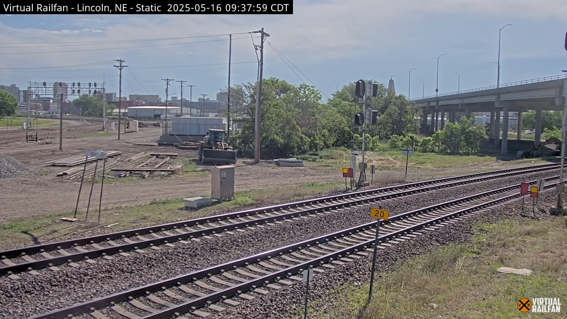 A view of railroad tracks under a partly cloudy sky, with construction equipment and materials visible in a nearby area, and a highway overpass in the background.