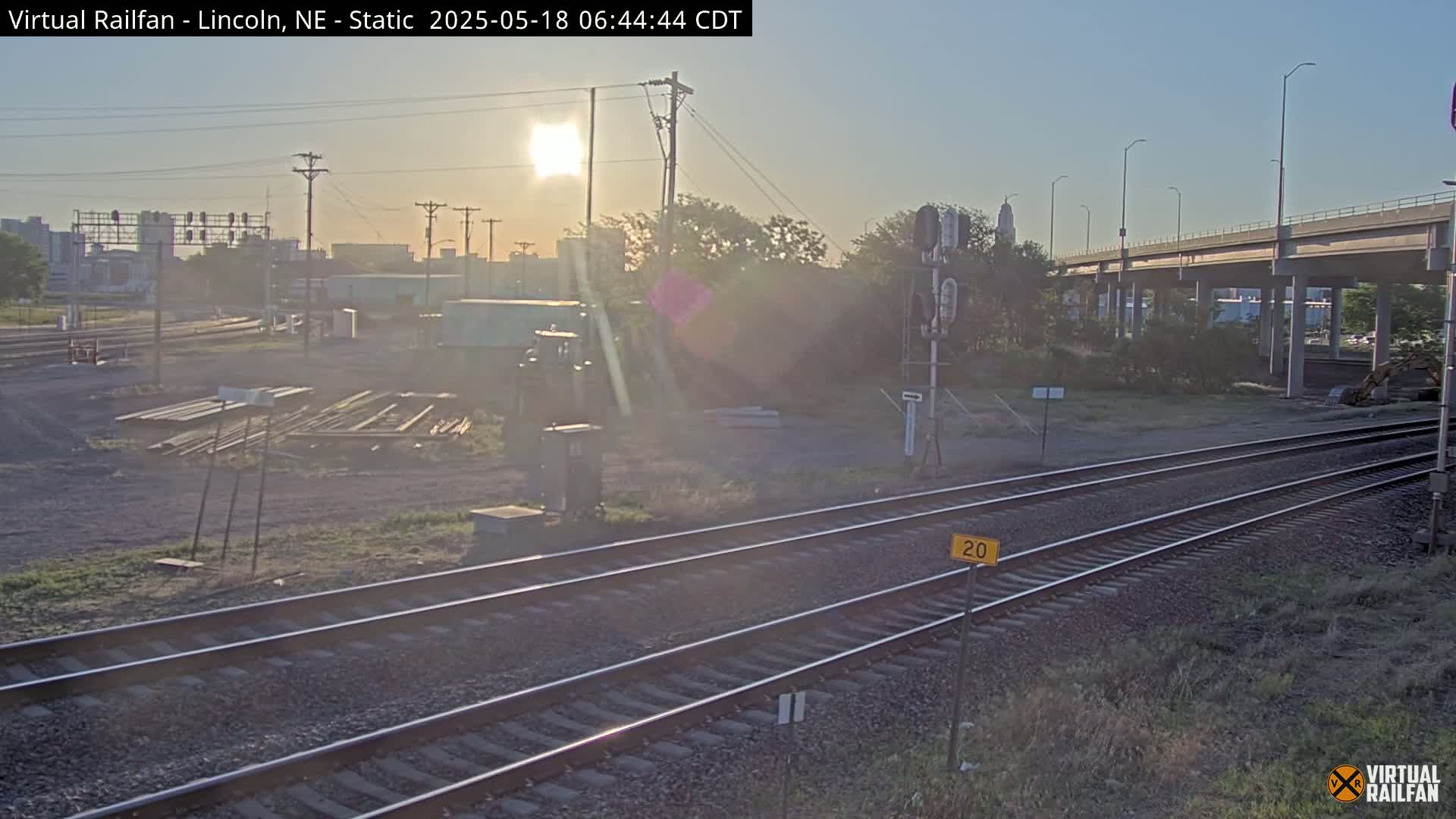 A sunrise view of railroad tracks and a highway overpass, with some industrial equipment and buildings in the background.