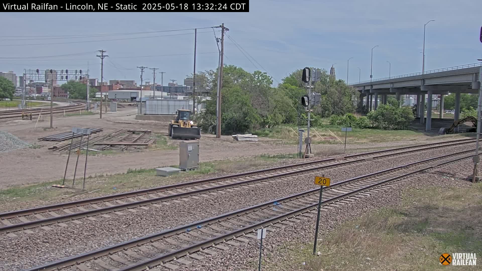 A sunny day view of railroad tracks undergoing construction near a highway overpass, with a yellow construction vehicle visible in the distance.