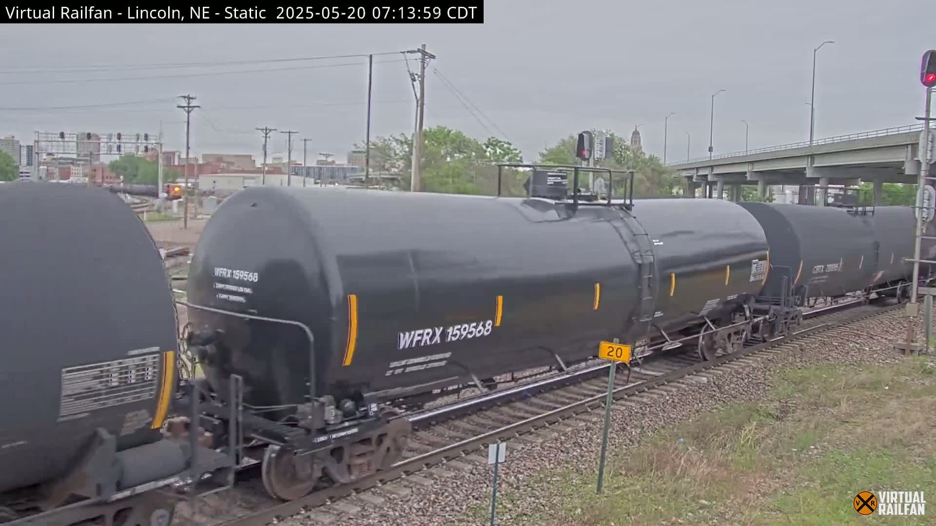 A long freight train of dark-colored tanker cars moves along railroad tracks under an overcast sky.