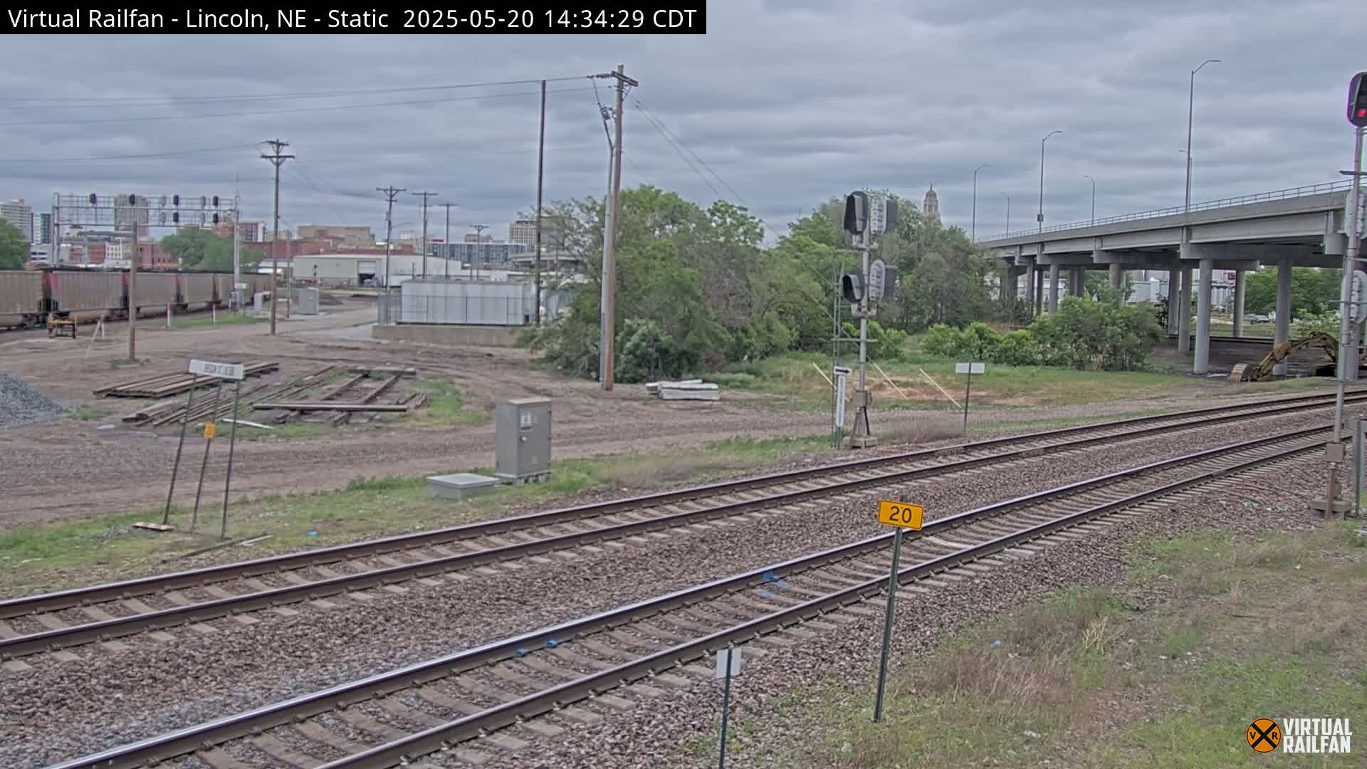 A view of railroad tracks under an overcast sky, with a distant cityscape and a highway overpass visible in the background.