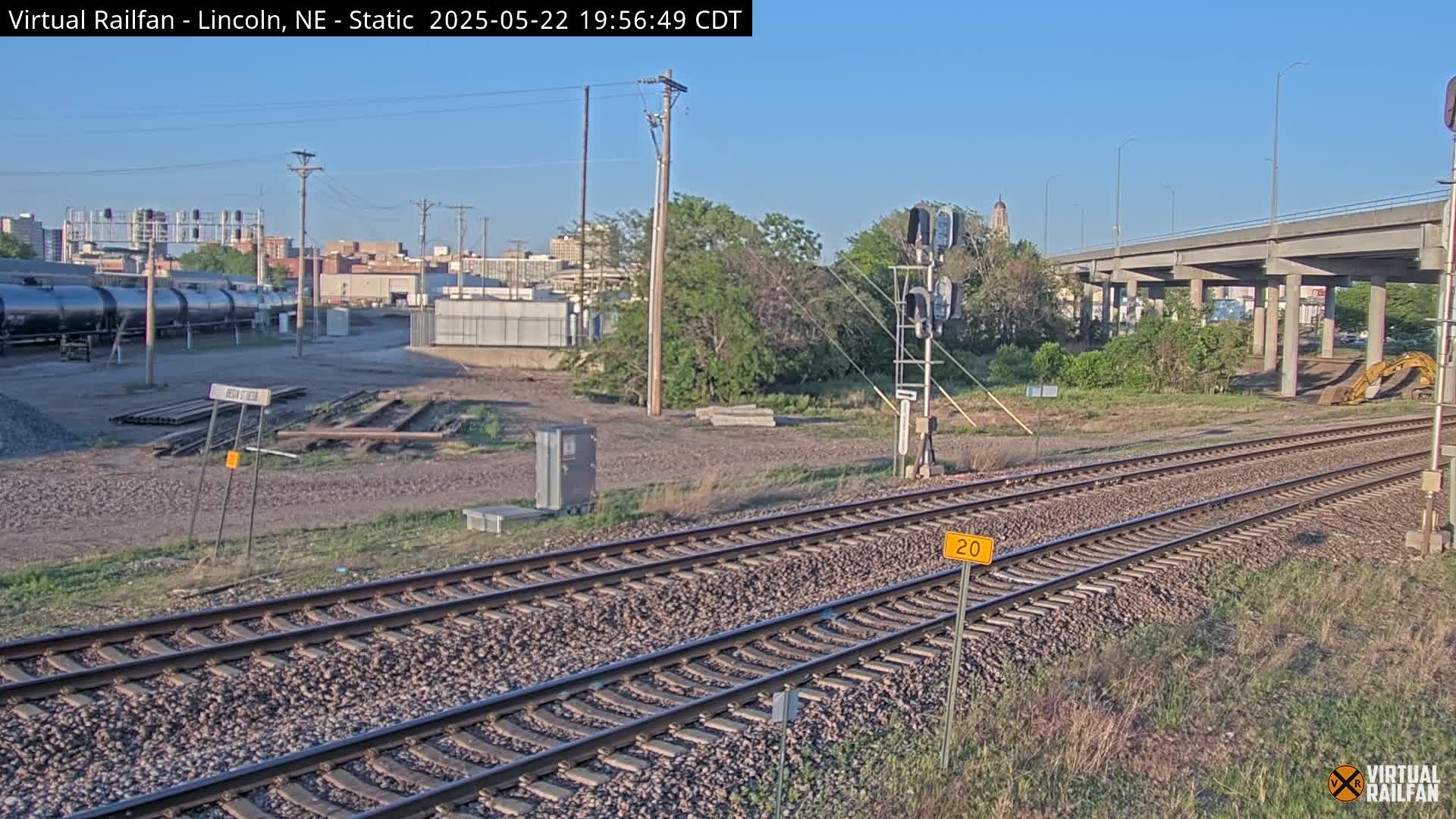 A section of railroad tracks under a clear sky, near a city skyline, with some light vegetation and a concrete overpass visible in the background.