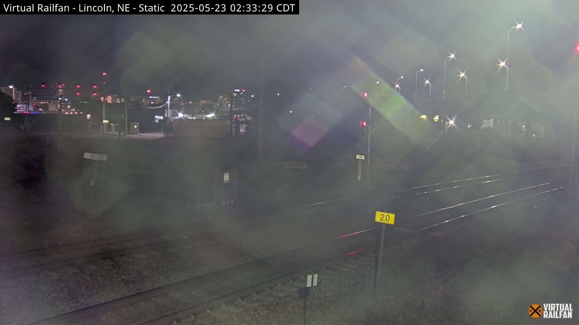 A nighttime view of railroad tracks under hazy conditions, with a city skyline visible in the distance.