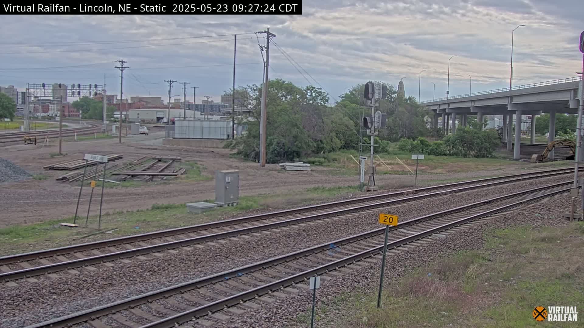 A view of railroad tracks under an overcast sky, with a highway overpass in the background and some construction equipment visible.