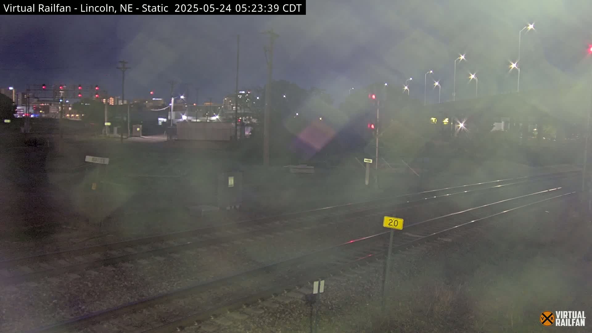 A nighttime view of railroad tracks under a hazy, dark sky, with city lights visible in the distance.