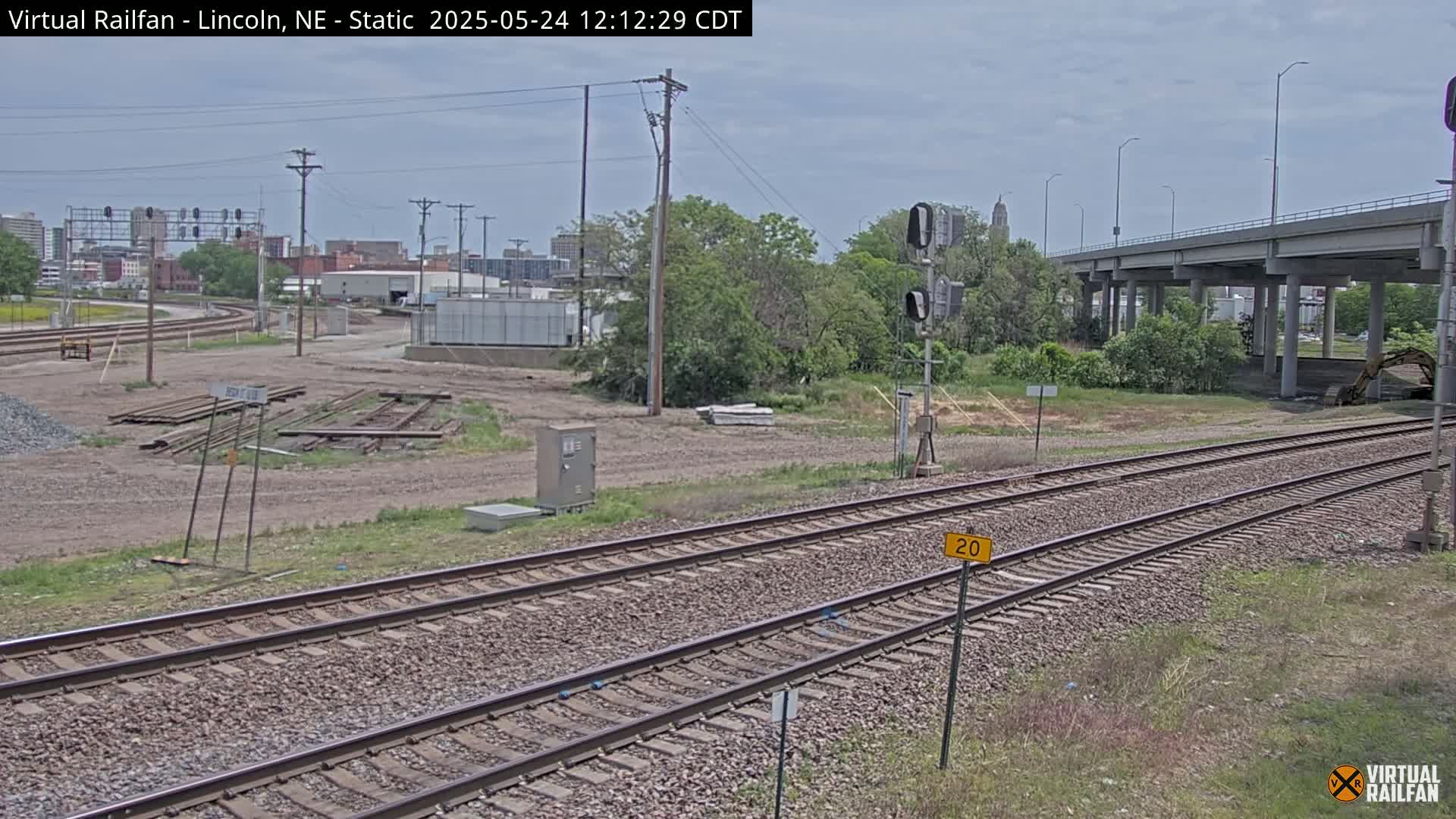 Two sets of railroad tracks extend into the distance under a partly cloudy sky, passing beneath an overpass near a city skyline and some construction equipment.