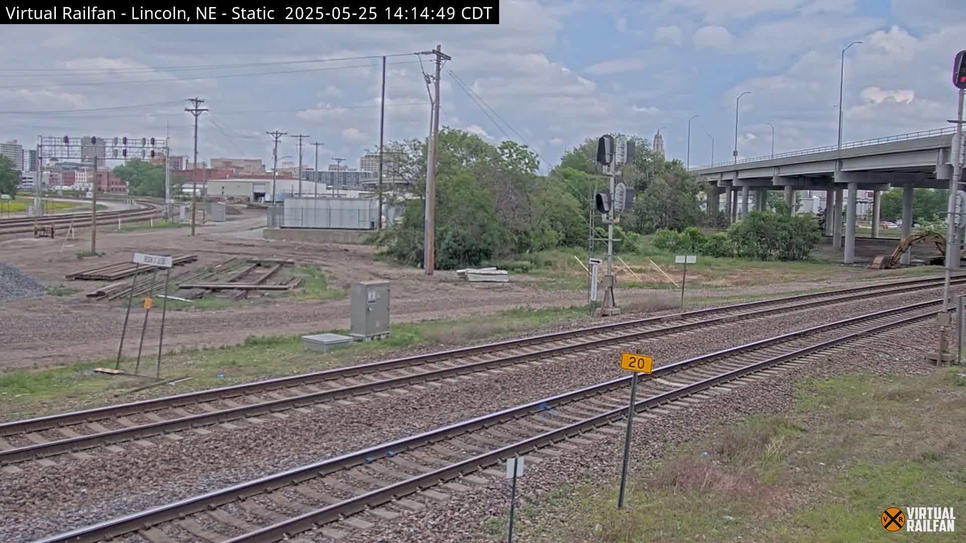 A partly cloudy day reveals railroad tracks in a construction area, with a highway overpass and distant cityscape in the background.