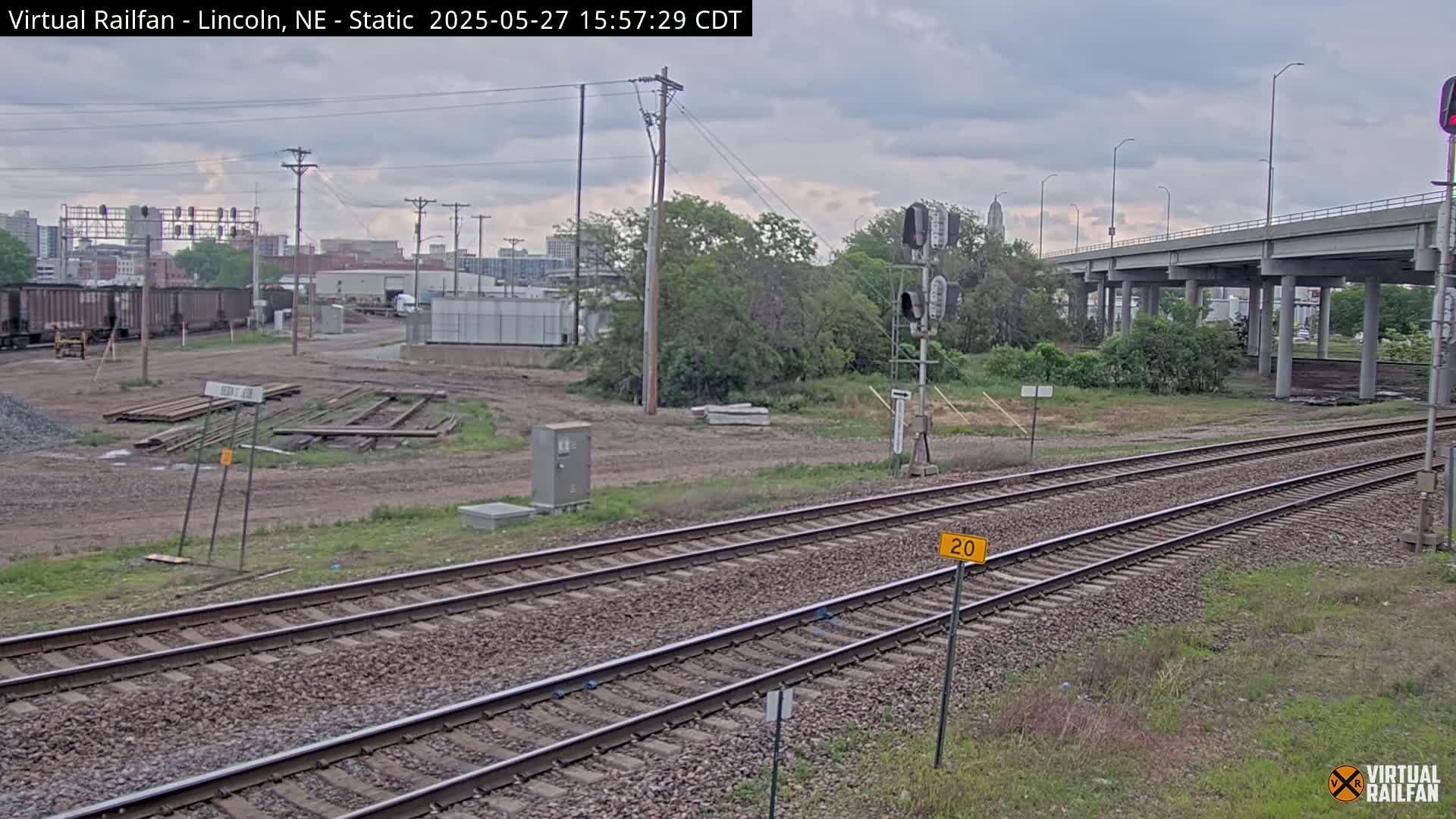 A view of railroad tracks under a cloudy sky, with a highway overpass in the background and some industrial buildings and vegetation visible.