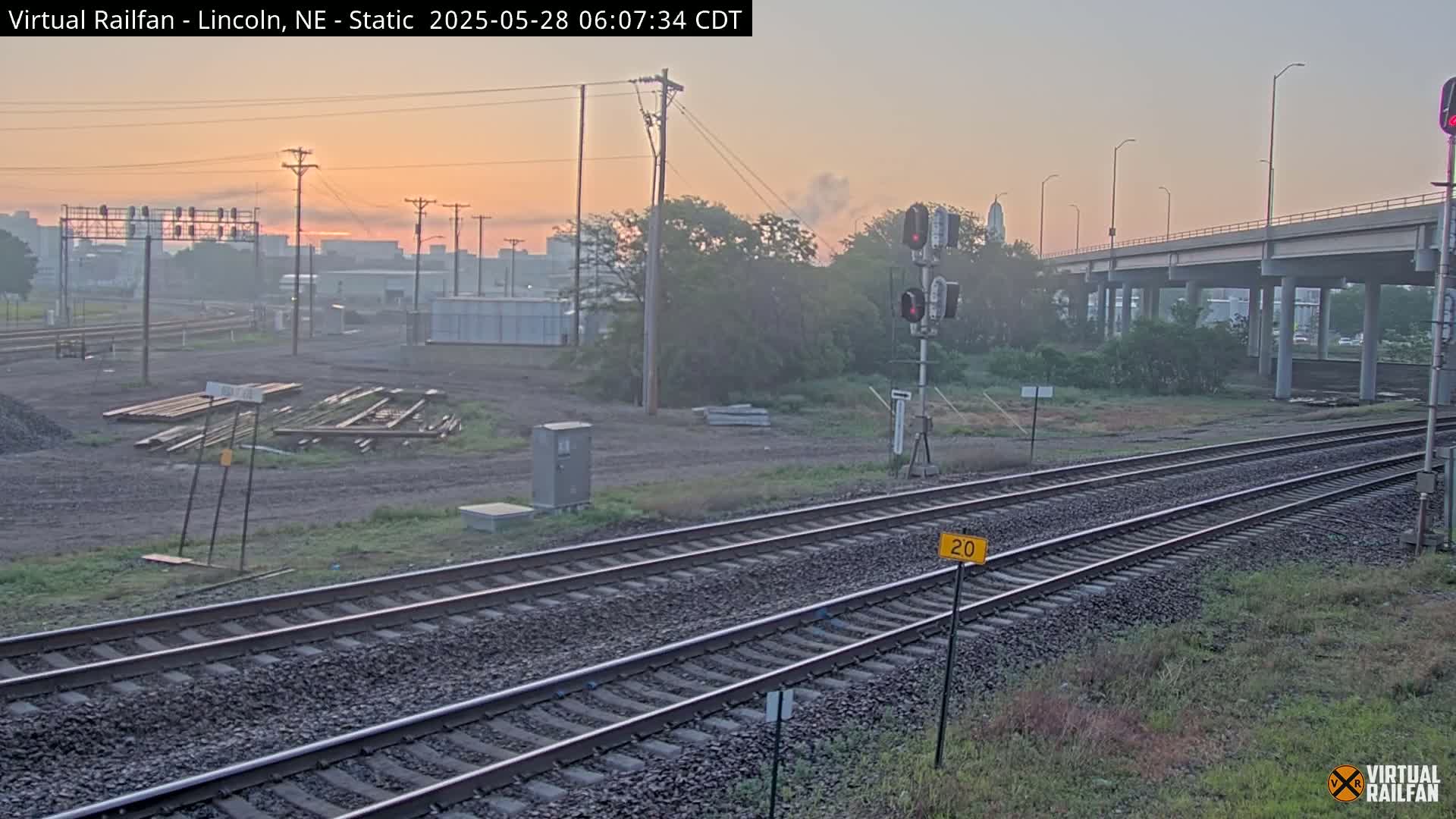 A hazy sunrise illuminates a railway scene with two sets of tracks, a highway overpass in the background, and some construction materials lying near the rails.