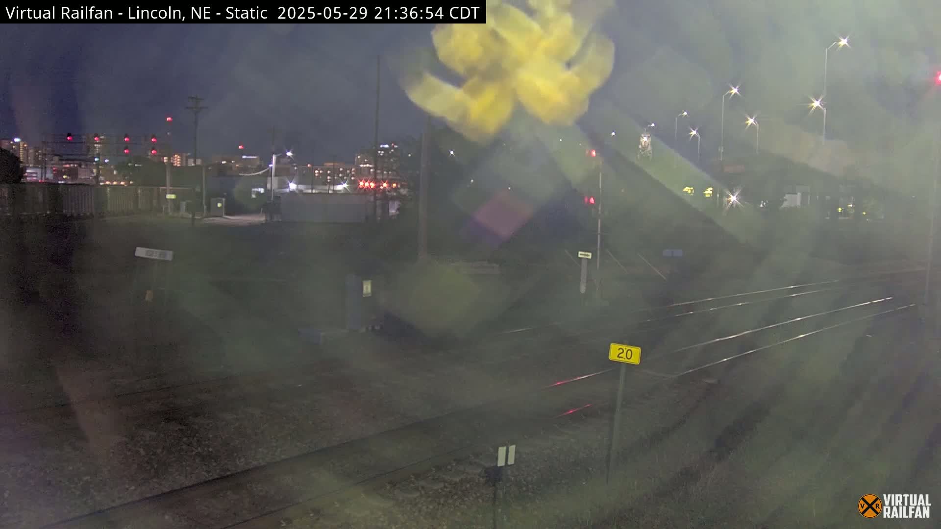 A nighttime view of railroad tracks and a city skyline in the distance, with hazy or rainy conditions obscuring the view.