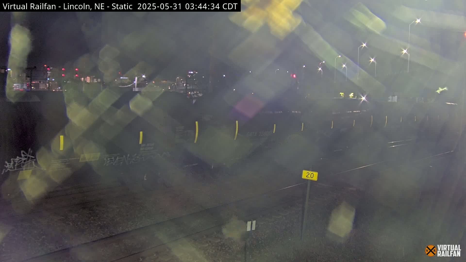 A nighttime view of a freight train on railroad tracks, under hazy conditions with city lights in the background.