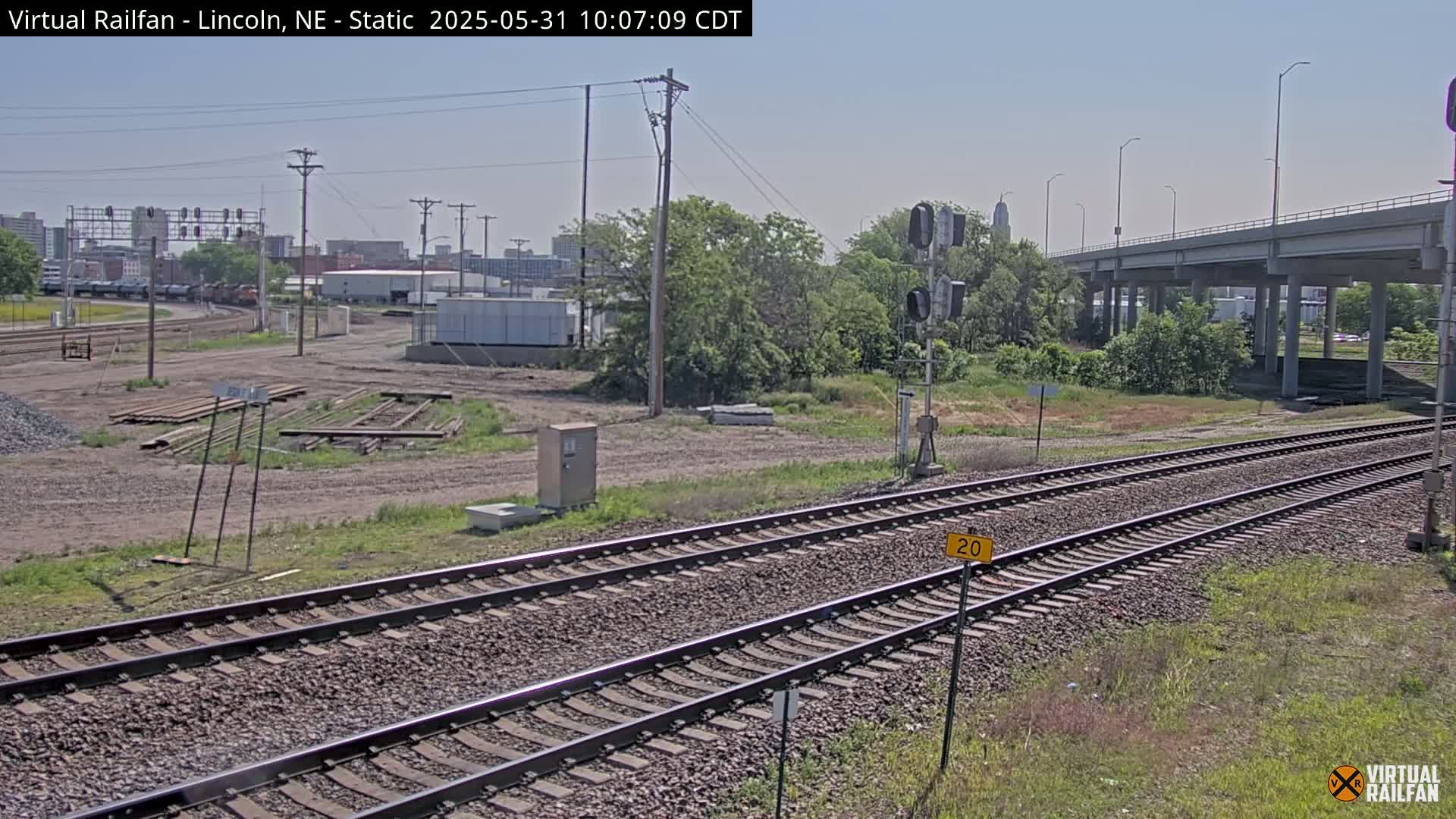 Under a clear sky, railroad tracks extend toward a city skyline, passing beneath a highway overpass amidst some vegetation and industrial areas.
