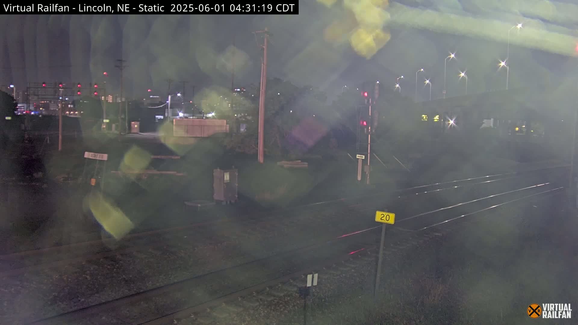 A nighttime view of a railroad crossing with hazy conditions, showing train tracks, a signal, and lights in the distance.